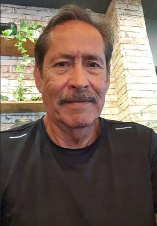 Hombre de edad media con bigote usando camisa negra, posando en interiores con pared de ladrillo y planta verde detrás