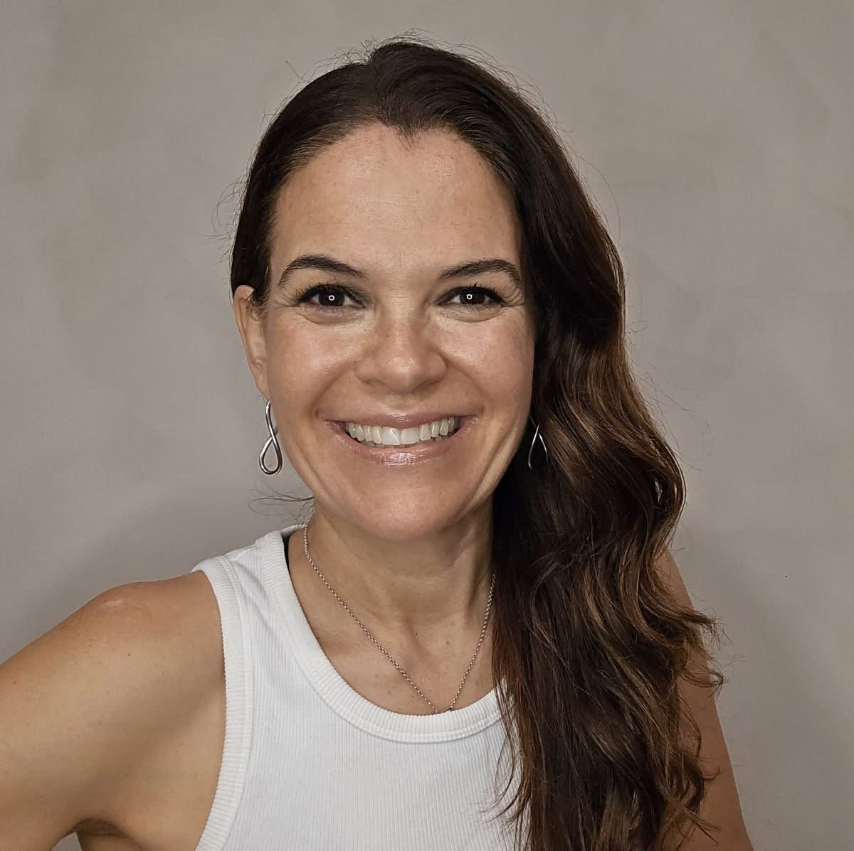Retrato de mujer sonriente con cabello oscuro ondulado largo, camiseta blanca sin mangas y aretes de aro sobre fondo gris neutro