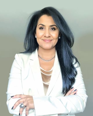 Professional headshot of a woman with long dark hair wearing a white blazer and layered necklace, smiling at camera with arms crossed