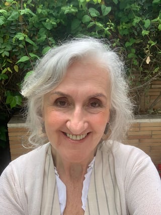 Smiling older woman with white hair wearing a beige cardigan outdoors with green foliage in background